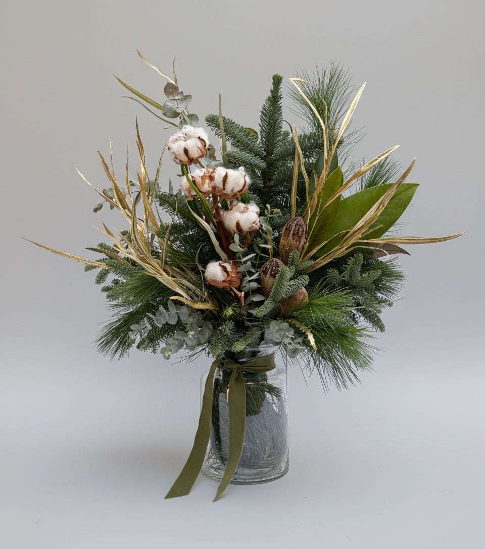 Ramo navideño de abeto y pino natural con una rama de algodón seco, lleva detales en dorados y va sobre un jarrón básico de cristal