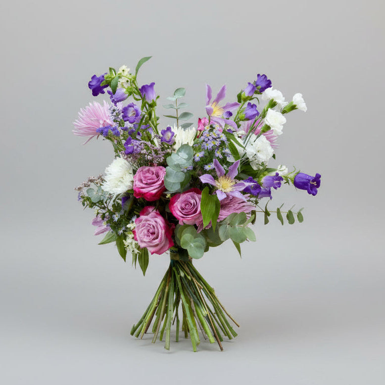 ramo en colores morados, malvas, rosas y blancos con toques verdes de eucalipto natural y atado con una cinta en color verde sobre un fondo gris claro con luz neutra