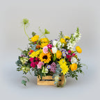 centro de flores naturales en colores primaverales con toques amarillos, naranjas, rosas  y verdes naturales montado sobre una caja de madera pequeña en natural.