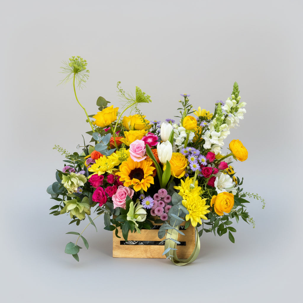 centro de flores naturales en colores primaverales con toques amarillos, naranjas, rosas  y verdes naturales montado sobre una caja de madera pequeña en natural.