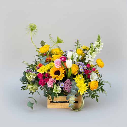 centro de flores naturales en colores primaverales con toques amarillos, naranjas, rosas  y verdes naturales montado sobre una caja de madera pequeña en natural.