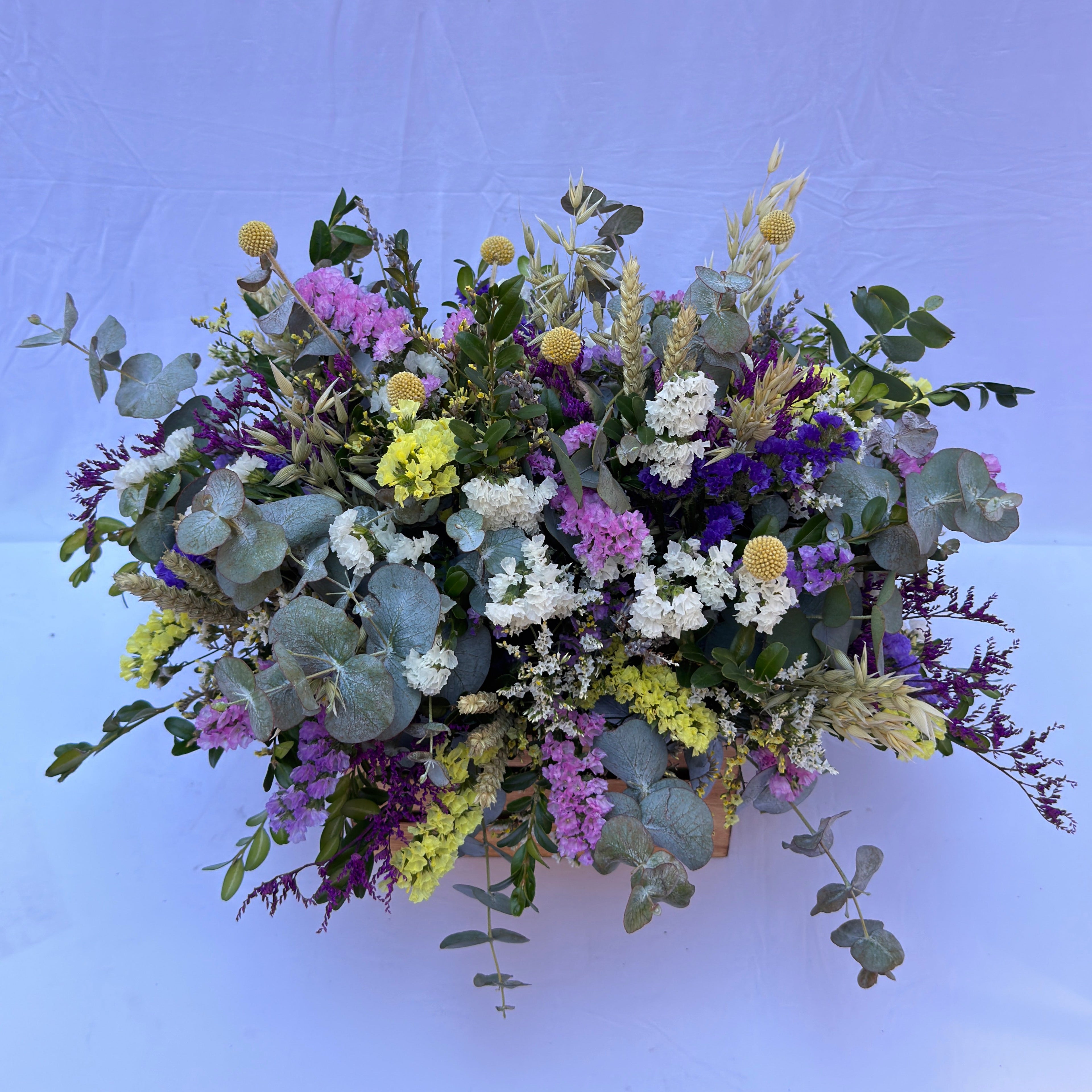 centro de flores secas colorido  sobre una caja de madera en color neutro, compuesto de estatis colorido, espiga natural y flores de campo en seco