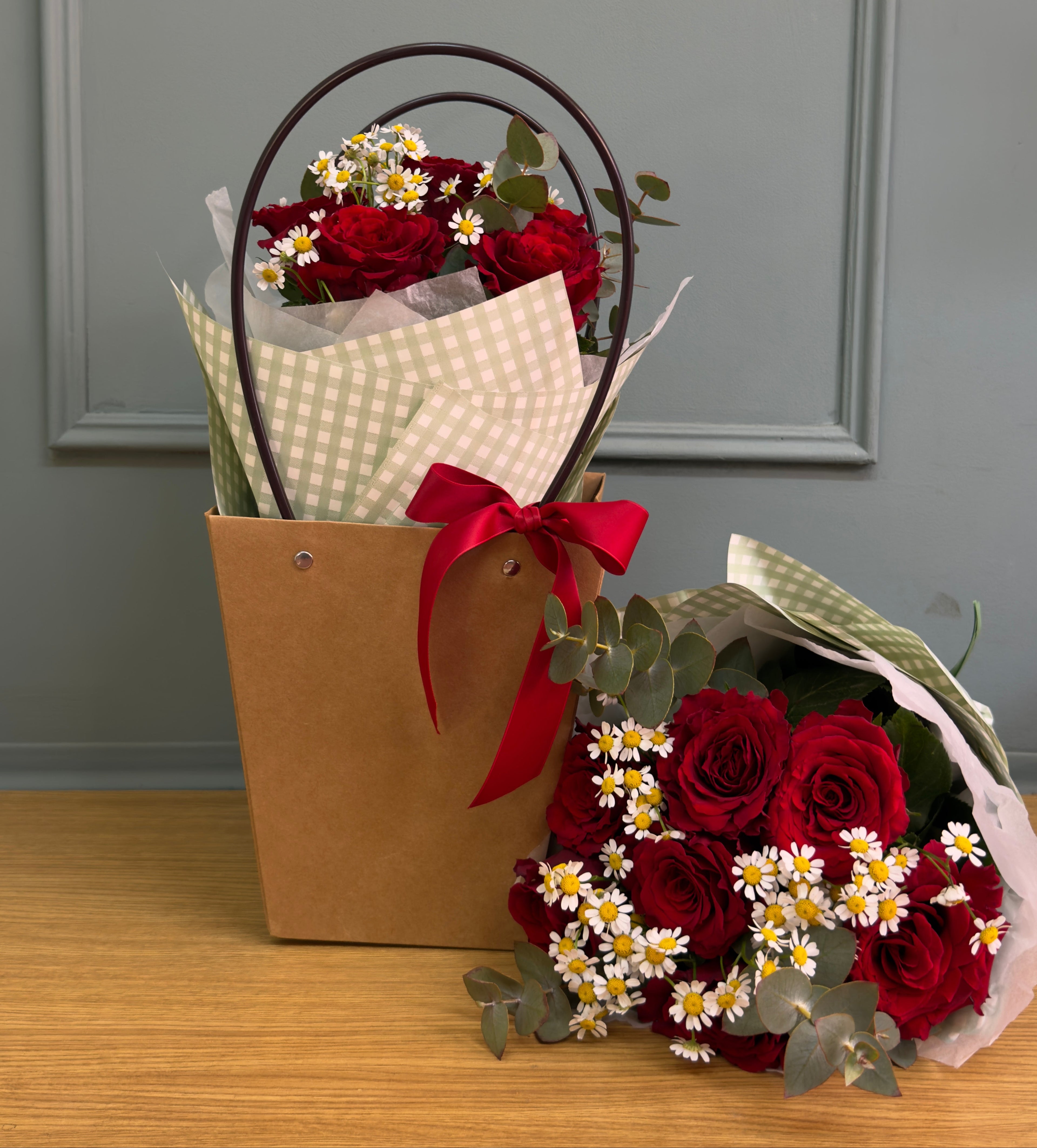 ramo en forma de bouquet compacto pequeño con flores naturales de rosas rojas con toque de flor de manzanilla y verdes naturales. El ramo va sobre una caja de kraft y azas negras rematado por un lazo rojo