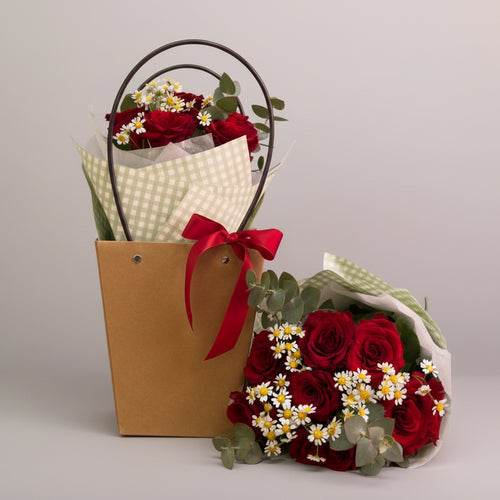 ramo en forma de bouquet compacto pequeño con flores naturales de rosas rojas con toque de flor de manzanilla y verdes naturales. El ramo va sobre una caja de kraft y azas negras rematado por un lazo rojo
