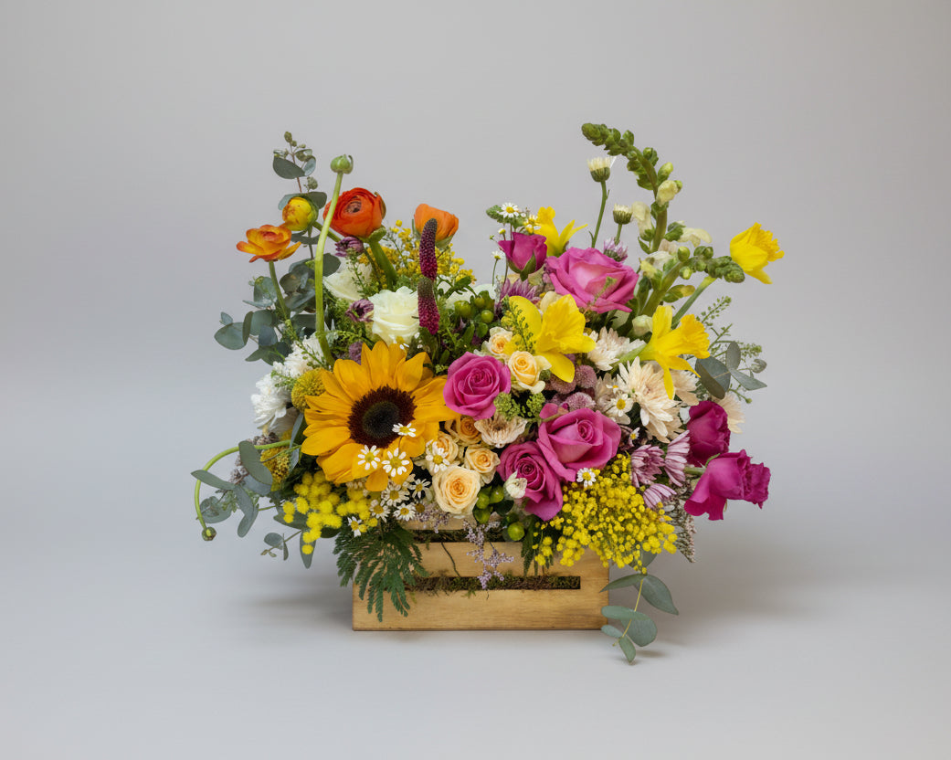 centro de flores naturales en colores primaverales con toques amarillos, naranjas, rosas  y verdes naturales montado sobre una caja de madera pequeña en natural.