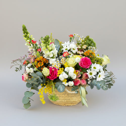 centro floral con flores de temporada, sobre un capazo de mimbre artesanal rematado por una cinta en verde de satén.