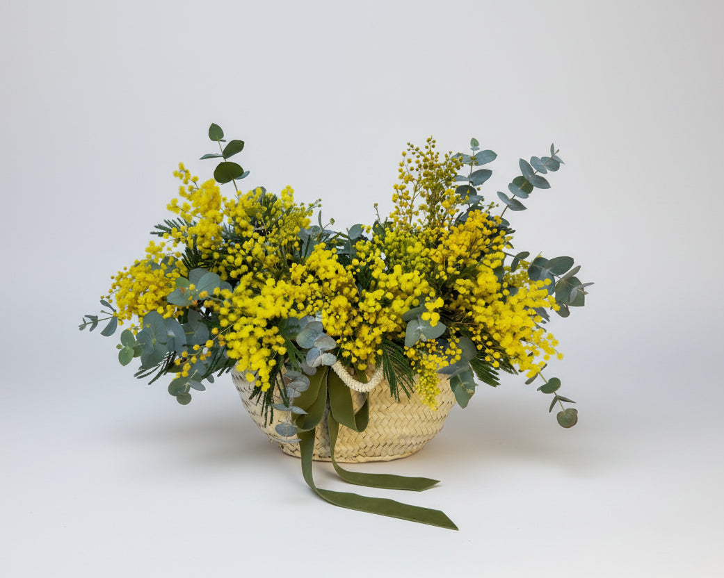 centro floral de mimosa y eucalipto natural sobre un capazo de rafia rematado por un lazo de terciopelo color verde botella