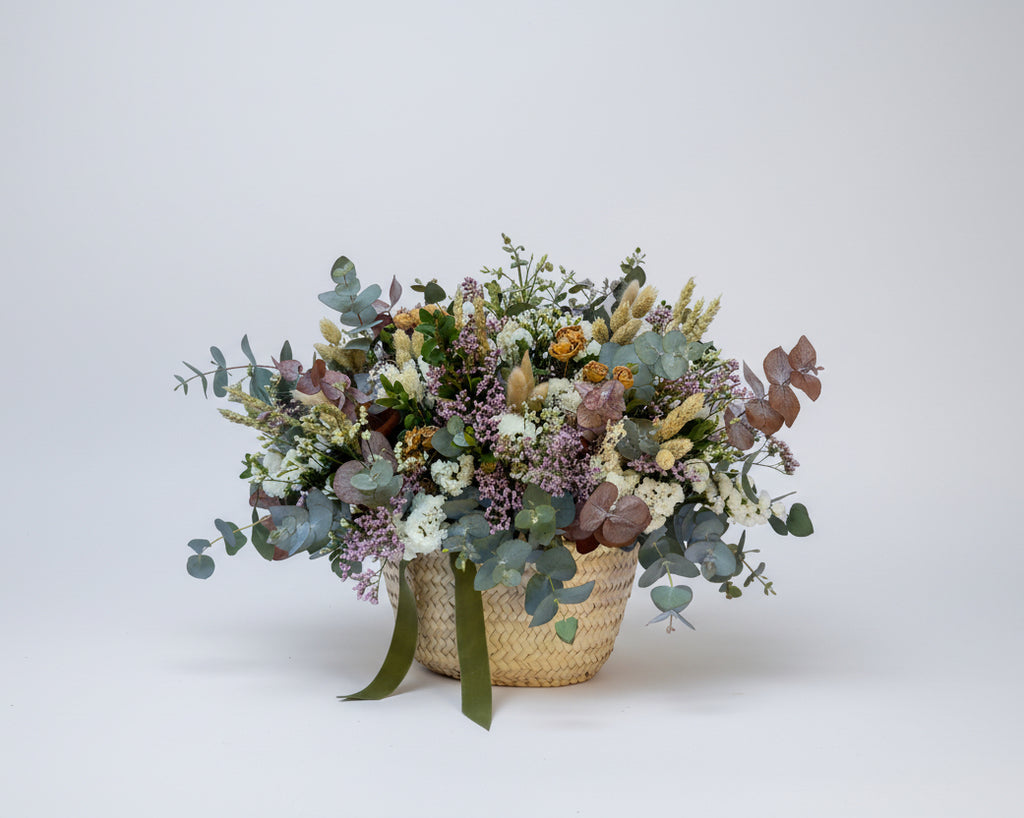 cesto de flores secas en color blancos y verdes de eucalipto y flores de campo preservadas sobre una base de mimbre beiges con cinta de terciopelo verde botella