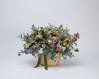 cesto de flores secas en color blancos y verdes de eucalipto y flores de campo preservadas sobre una base de mimbre beiges con cinta de terciopelo verde botella
