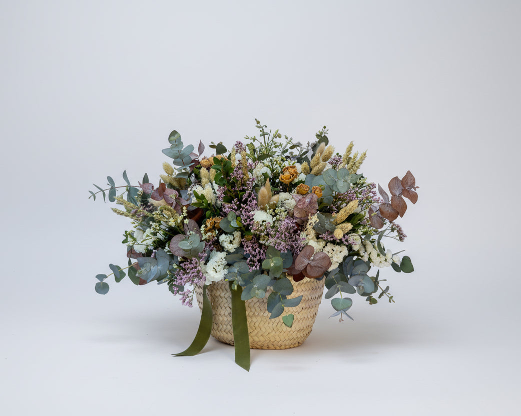 cesto de flores secas en color blancos y verdes de eucalipto y flores de campo preservadas sobre una base de mimbre beiges con cinta de terciopelo verde botella