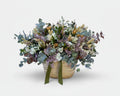 cesto de flores secas en color blancos y verdes de eucalipto y flores de campo preservadas sobre una base de mimbre beiges con cinta de terciopelo verde botella