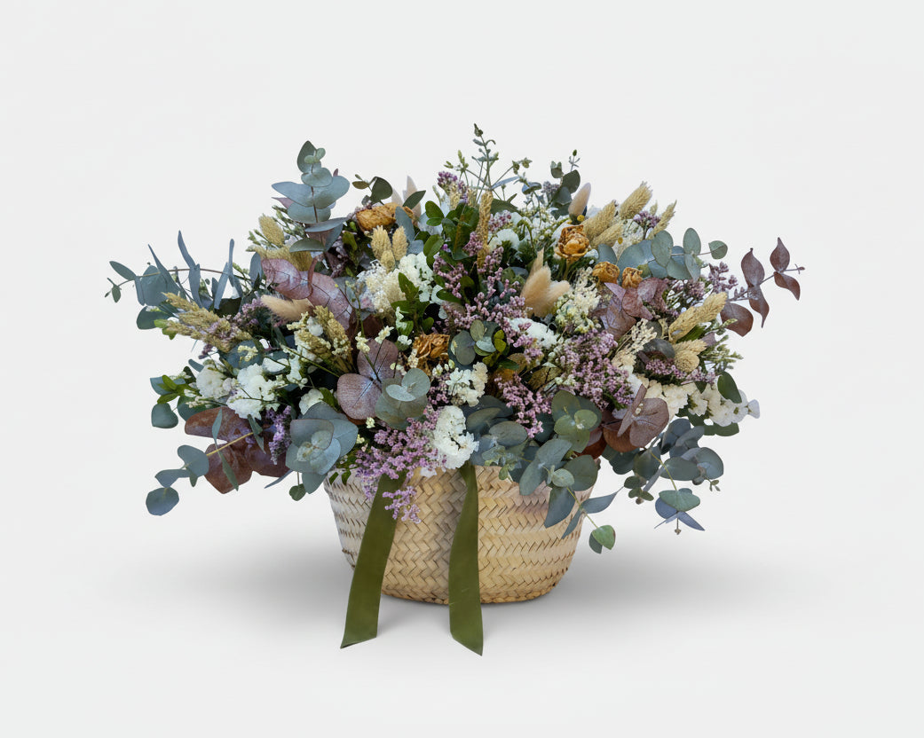 cesto de flores secas en color blancos y verdes de eucalipto y flores de campo preservadas sobre una base de mimbre beiges con cinta de terciopelo verde botella