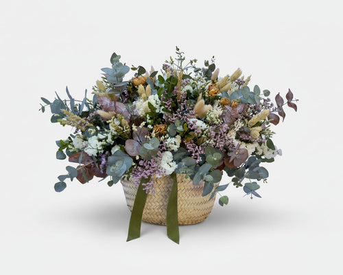 cesto de flores secas en color blancos y verdes de eucalipto y flores de campo preservadas sobre una base de mimbre beiges con cinta de terciopelo verde botella