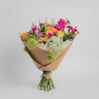 en la imagen se muestra un ramo de flores primaverales en color rosa fucsia, color salmón, rosas y verdes naturales de eucalipto envuelto con papel kraft y atado con rafia color verde en un fondo gris claro y luz neutra.