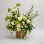 centro de flores naturales con rosas, crisantemos y margaritas blancas con toques despuntado de delfinium azul claro, también tiene toques heleborus en verdes y blancos, el centro va dentro de un macetero marron de yute artesanal y tocado con un lazo verde, toda la composición está en un fondo blanco limpio.