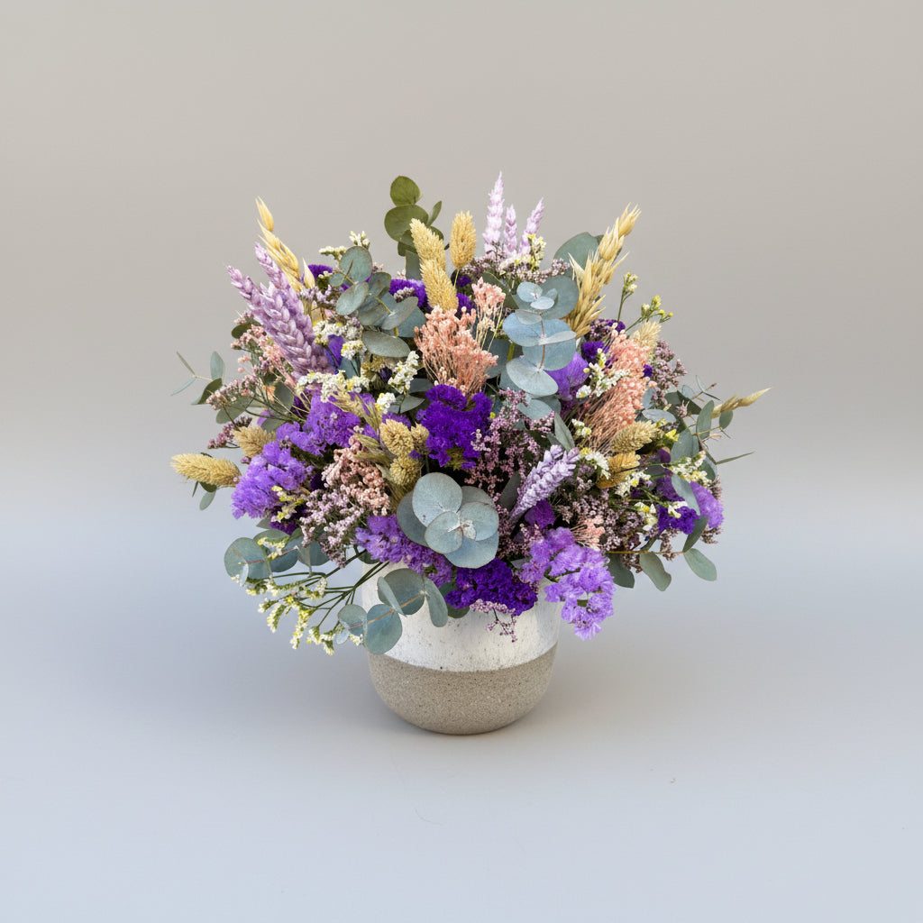 centro de flores secas en colores morados y lilas con toque de verdes naturales sobre base de ceramica bicolor blanco y gris.