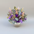 centro de flores secas en colores morados y lilas con toque de verdes naturales sobre base de ceramica bicolor blanco y gris.