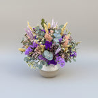 centro de flores secas en colores morados y lilas con toque de verdes naturales sobre base de ceramica bicolor blanco y gris.