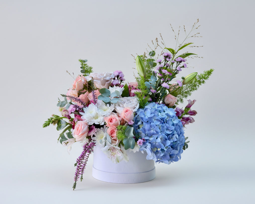 Centro de flores naturales en tonos claros y verdes sobre una base de cartón estilo sombrerera. 