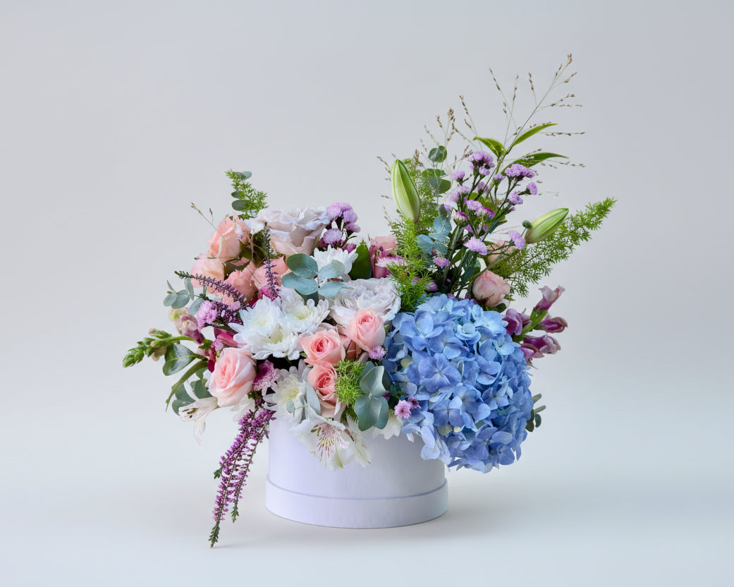 Centro de flores naturales en tonos claros y verdes sobre una base de cartón estilo sombrerera. 