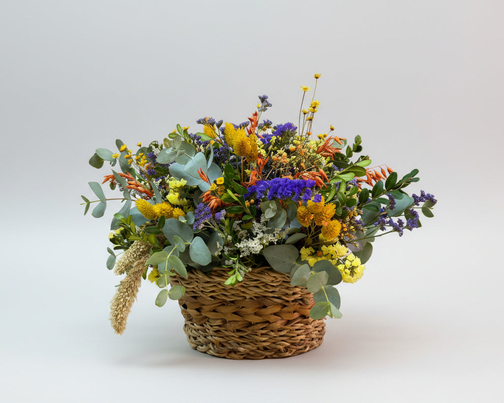 cesto de flores secas coloridas en tonos morados, mostaza y naranja, complementado con eucalipto natural, sobre un cesto de yute marrón