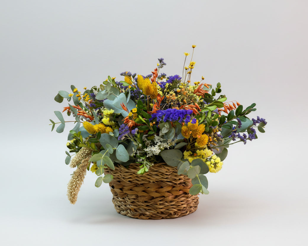 cesto de flores secas coloridas en tonos morados, mostaza y naranja, complementado con eucalipto natural, sobre un cesto de yute marrón