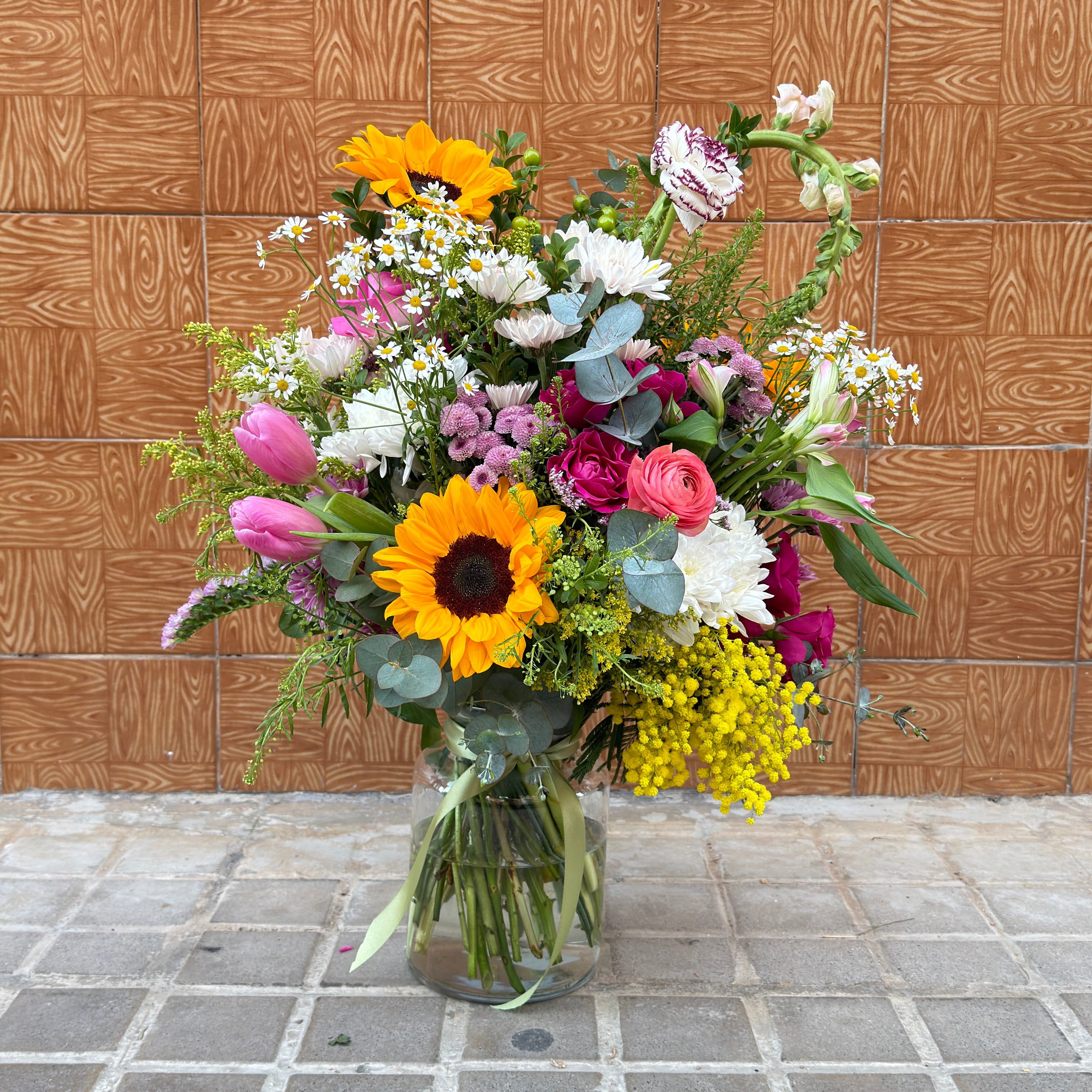 ramo de colores primaverales sobre jarrón transparente y rematado por un lazo verde de satén.