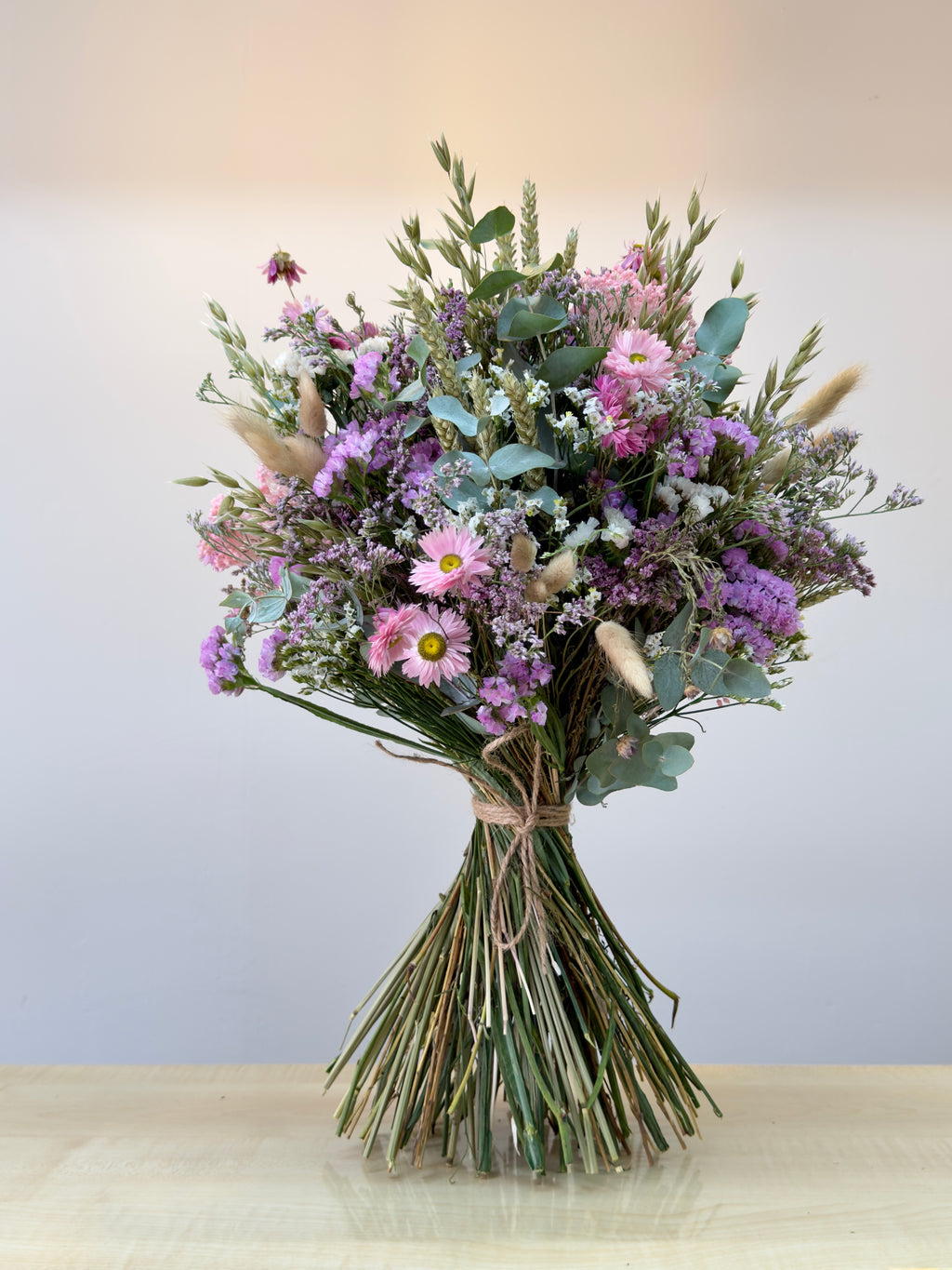ramo de flores secas en color rosa, blancos y verdes, compuesto por flores de siemprevivas, limonium y toques de espiga y cebada natural con flores de campo en seco. el ramo está atado con una cuerda de yute marrón con sus tallos limpios. toda la imagen está sobre un fondo blanco en luz neutra.