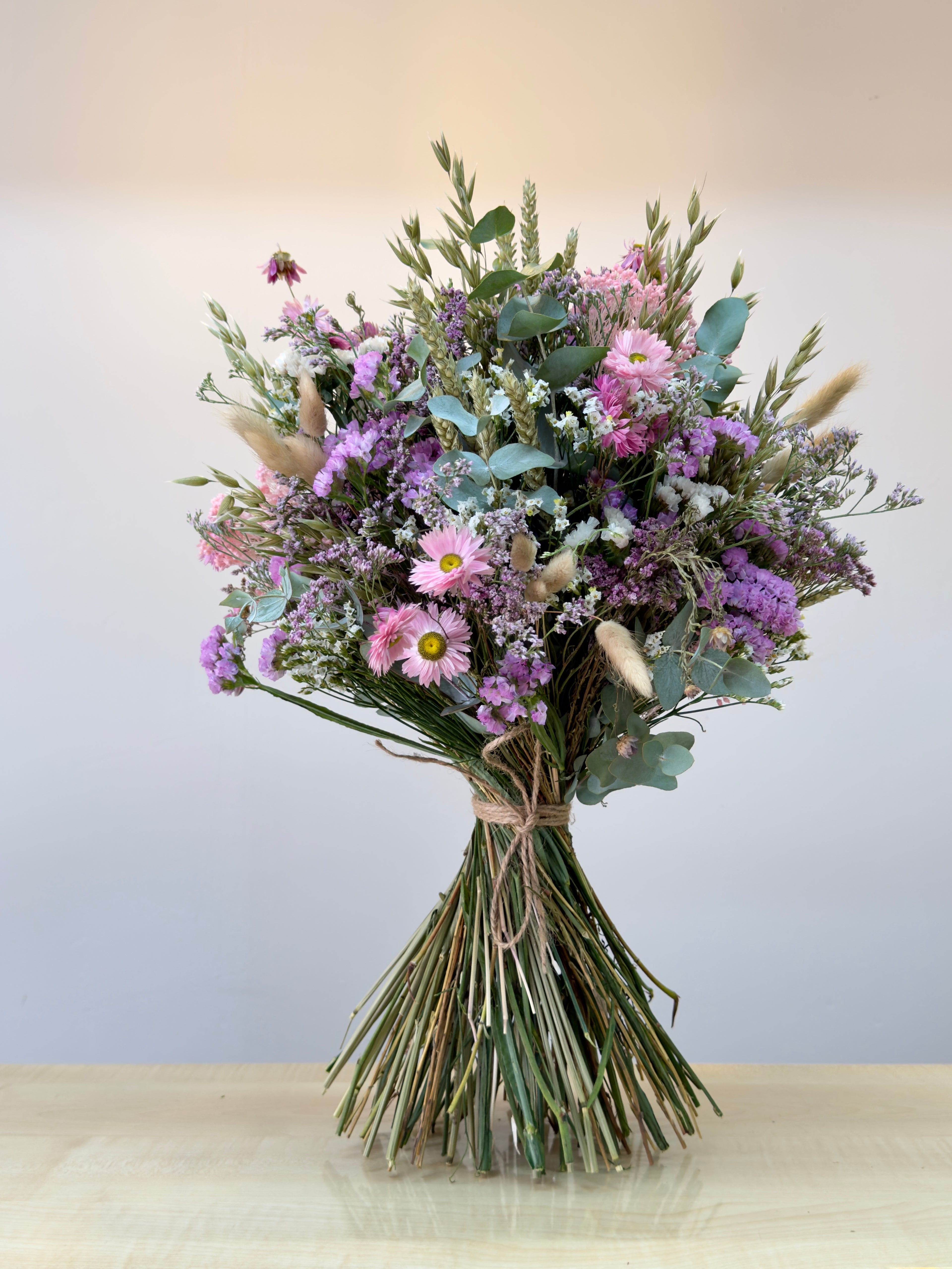 ramo de flores secas en color rosa, blancos y verdes, compuesto por flores de siemprevivas, limonium y toques de espiga y cebada natural con flores de campo en seco. el ramo está atado con una cuerda de yute marrón con sus tallos limpios. toda la imagen está sobre un fondo blanco en luz neutra.