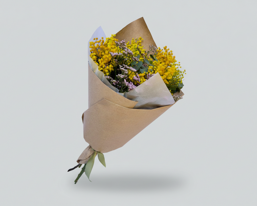 ramo de mimosa natural con toques de limonium rosa y eucalipto natural envuelto en papel seda blanco y papel kraft marrón atado con una cinta en color verde, todo el conjunto sobre un fondo blanco.