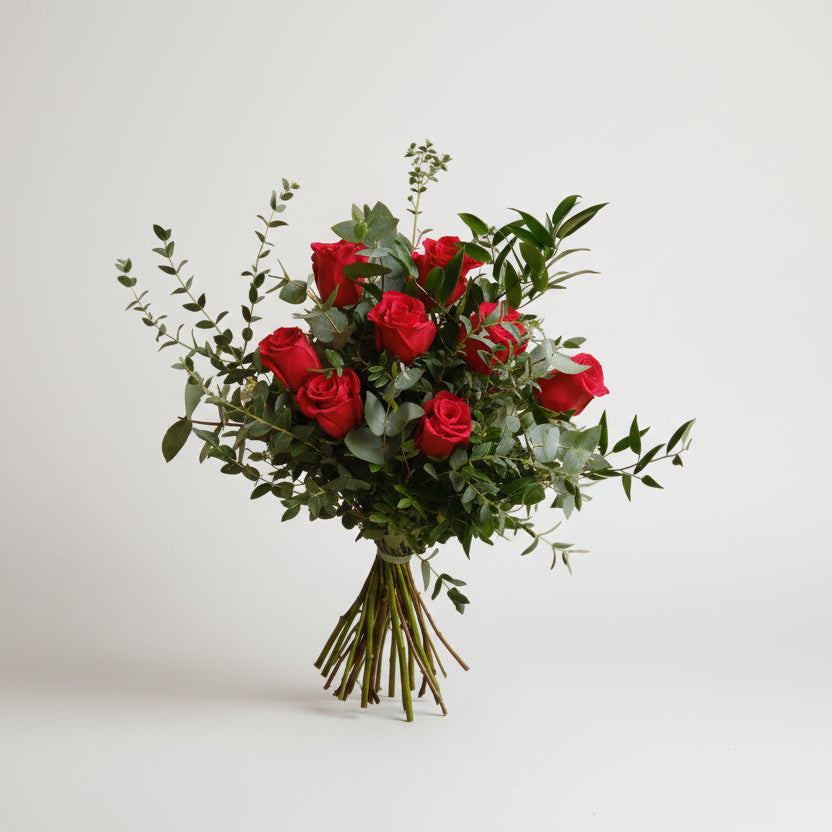 ramo de rosas rojas acompañado de follaje natural en distintas texturas como verdes de eucalipto, atado con una cita verde, todo el conjunto está sobre un fondo blanco.