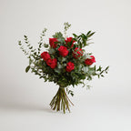 ramo de rosas rojas acompañado de follaje natural en distintas texturas como verdes de eucalipto, atado con una cita verde, todo el conjunto está sobre un fondo blanco.