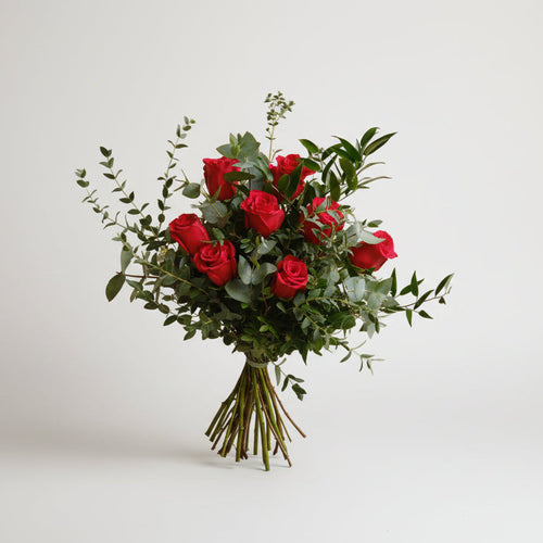 ramo de rosas rojas acompañado de follaje natural en distintas texturas como verdes de eucalipto, atado con una cita verde, todo el conjunto está sobre un fondo blanco.