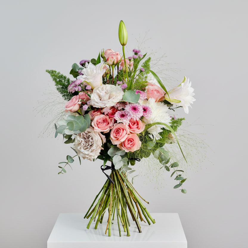 ramo en colores rosas, blancos y malvas tonalidad suave con toqus de verdes natural, atado con una cinta de afia. el ramo está sobre un pedestal blanco cuadrado en un fondo blanco