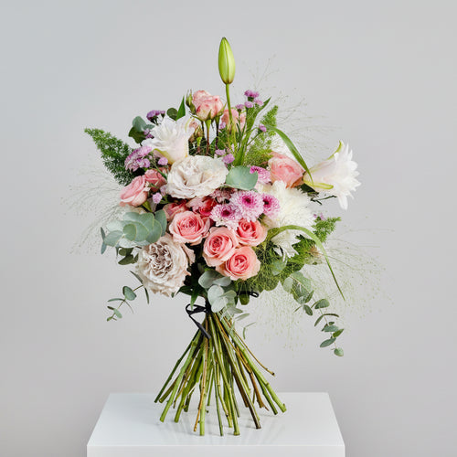 ramo en colores rosas, blancos y malvas tonalidad suave con toqus de verdes natural, atado con una cinta de afia. el ramo está sobre un pedestal blanco cuadrado en un fondo blanco